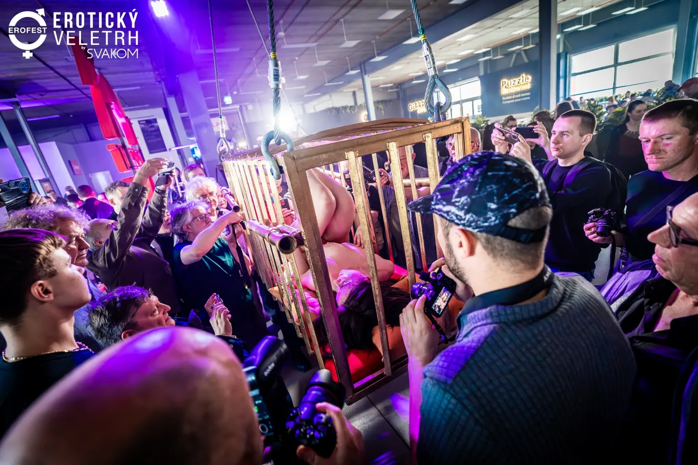 Diváci s fotoaparáty zblízka obklopují klec s performerkou ve speciální zóně